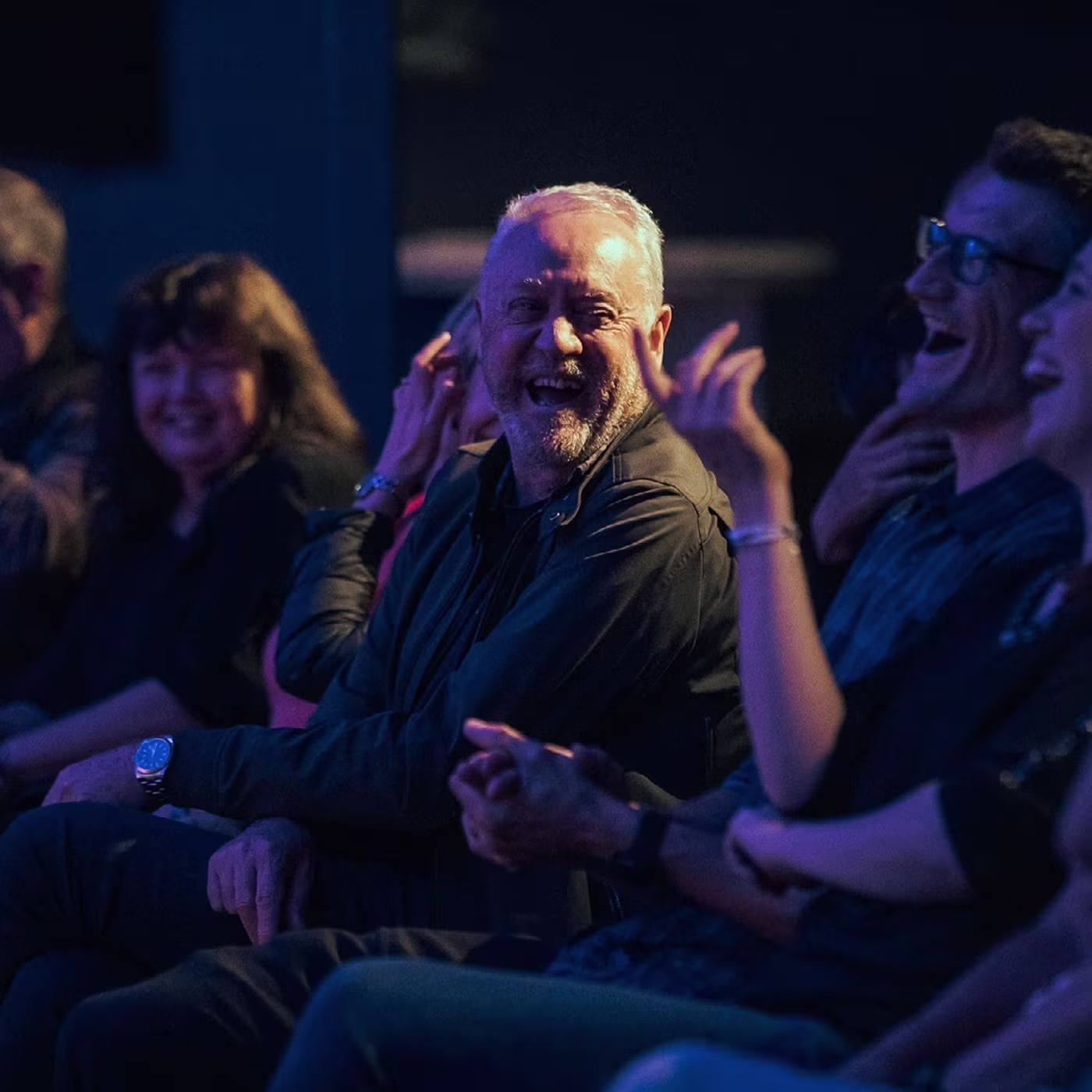 Audience members laughing and smiling while watching a live performance in a dimly lit venue.