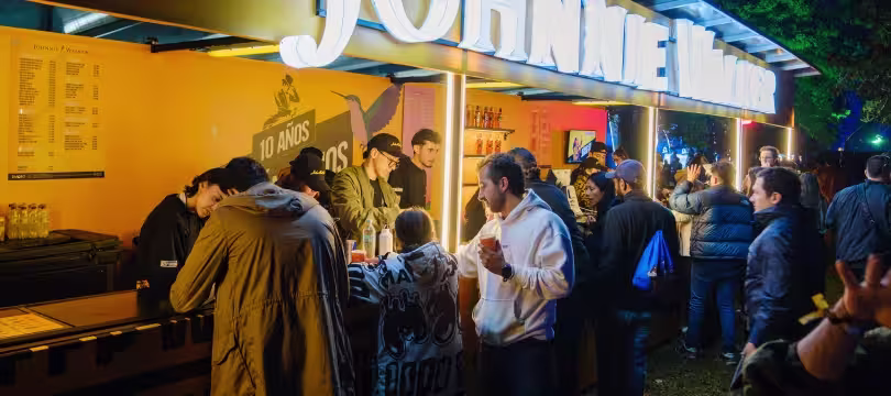 Una multitud se reúne en un bar Johnnie Walker al aire libre, bien iluminado, pidiendo bebidas y charlando. El personal atiende a los clientes tras el mostrador. El ambiente es animado y social, con árboles visibles al fondo.