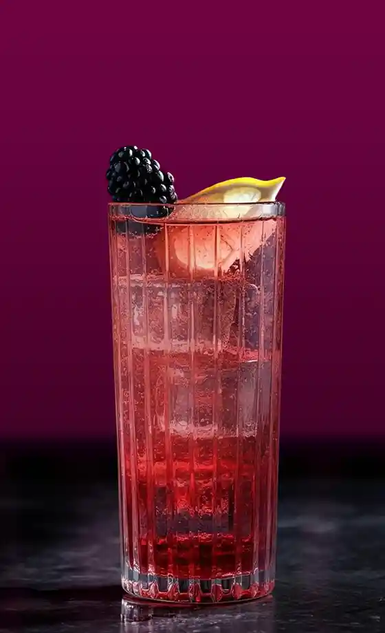 A tall glass filled with an effervescent red drink, topped with ice cubes. A slice of lemon and a blackberry decorate the drink. The background features deep purple drapes, adding an elegant touch.