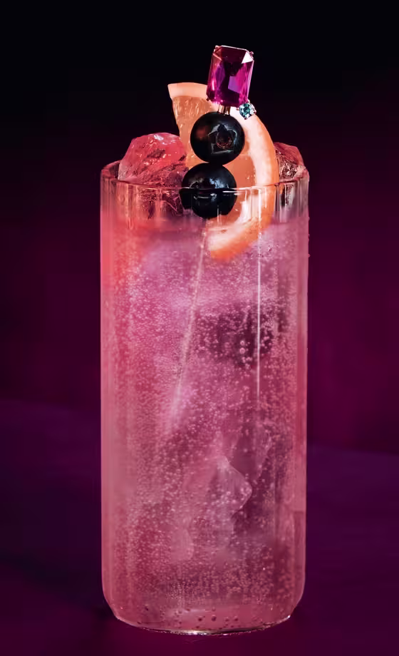 A tall glass filled with a sparkling pink cocktail, ice cubes, and garnished with two dark cherries and a grapefruit slice, sits on a magenta surface against a dark, gradient background.