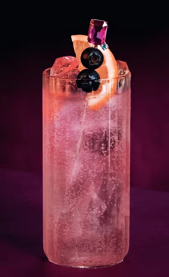 A tall glass filled with a sparkling pink cocktail, ice cubes, and garnished with two dark cherries and a grapefruit slice, sits on a magenta surface against a dark, gradient background.
