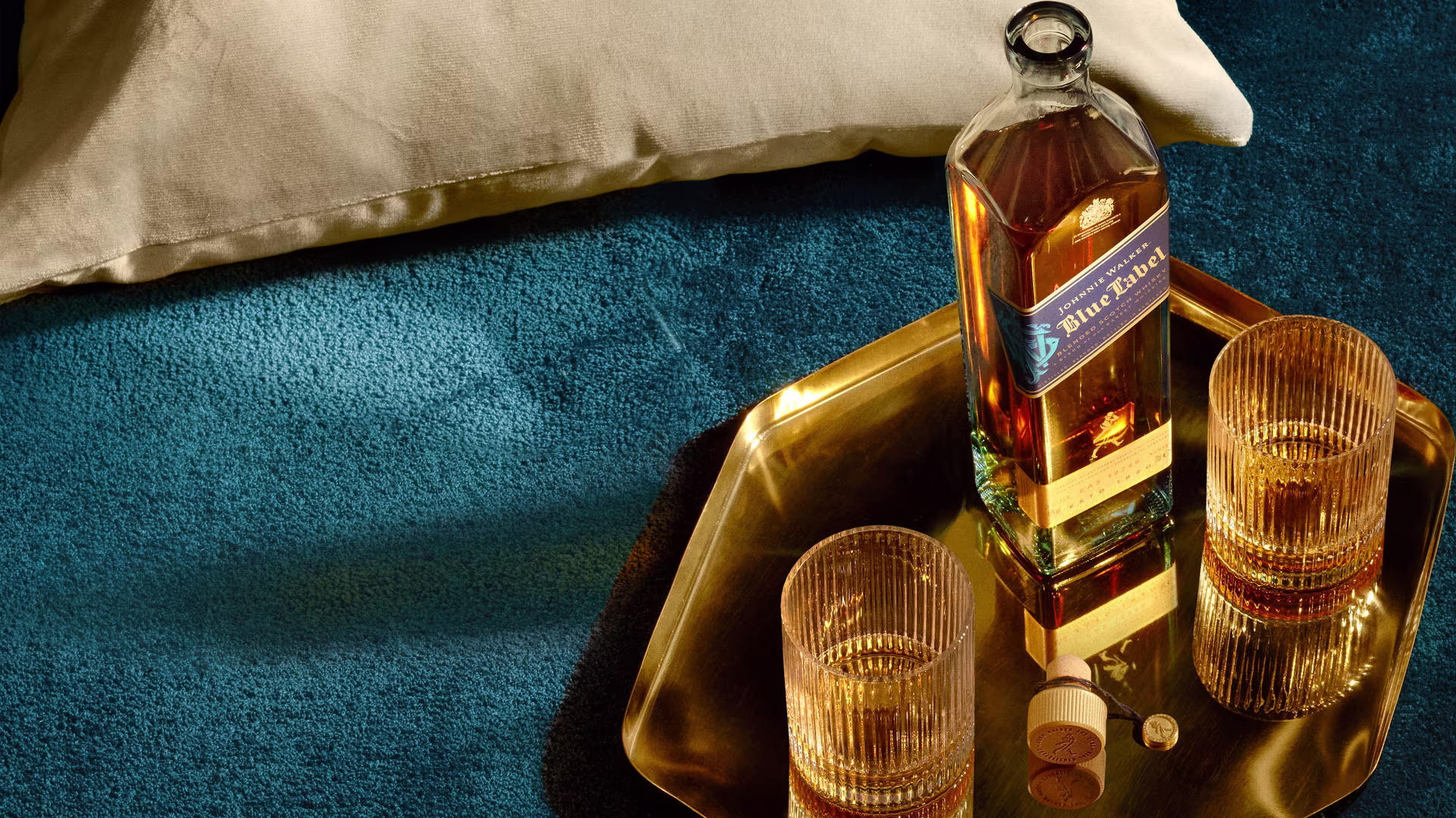 A bottle of Blue Label whiskey on a gold tray with two filled glasses and a cork, placed on a blue textured surface. A beige pillow is partially visible in the top left corner.