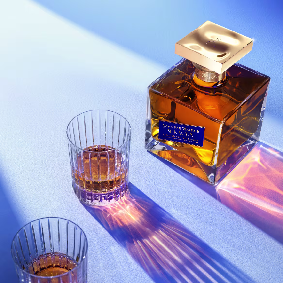 A bottle of Johnnie Walker Vault beside two crystal tumblers of whisky, lit in blue and amber light.