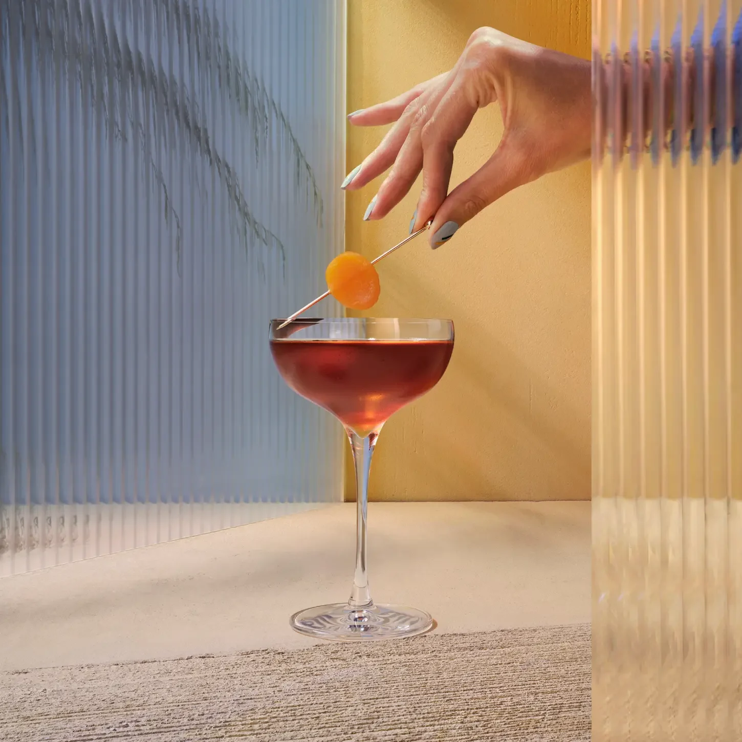 Le Old Fashioned Doux et Floral. Une main tient délicatement une brochette garnie d'orange au-dessus d'un cocktail servi dans un verre à pied. Le décor est composé de panneaux de verre texturés projetant des ombres sur un fond neutre et chaleureux.