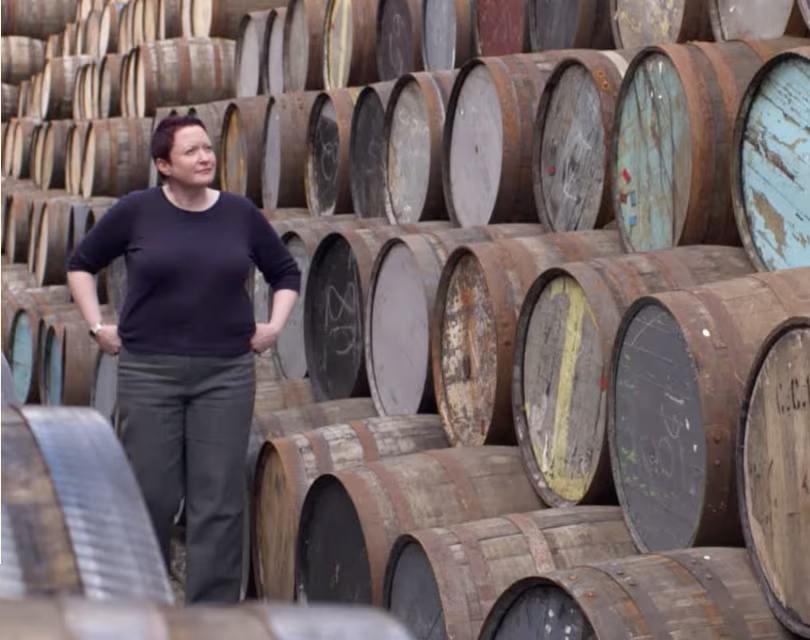 Une femme aux cheveux courts et portant un chandail foncé marche à côté de hautes piles de barils en bois patinés, certains avec de la peinture délavée, disposés les uns à côté des autres à l'extérieur.