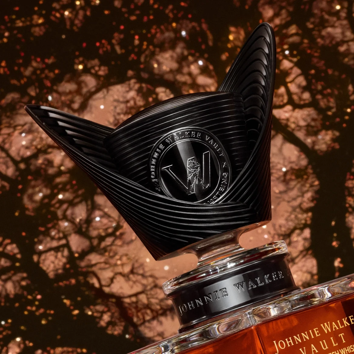 A close-up of a decorative Johnnie Walker whisky bottle stopper with a black, wing-like design, set against a blurred background of tree branches and glowing lights.