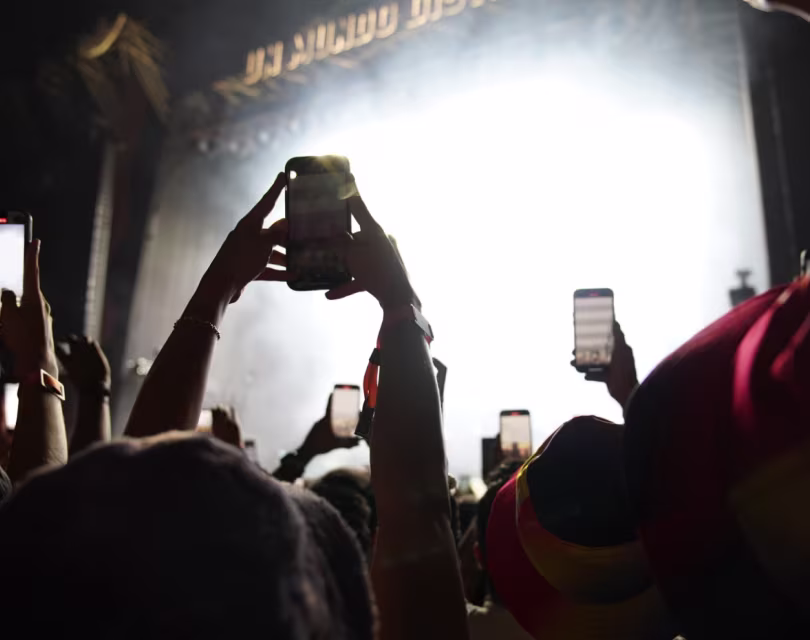 En un concierto, el público levanta sus teléfonos para grabar y capturar el escenario brillantemente iluminado. El fondo está iluminado por una intensa luz blanca, y algunas personas llevan sombreros coloridos, creando un ambiente animado.