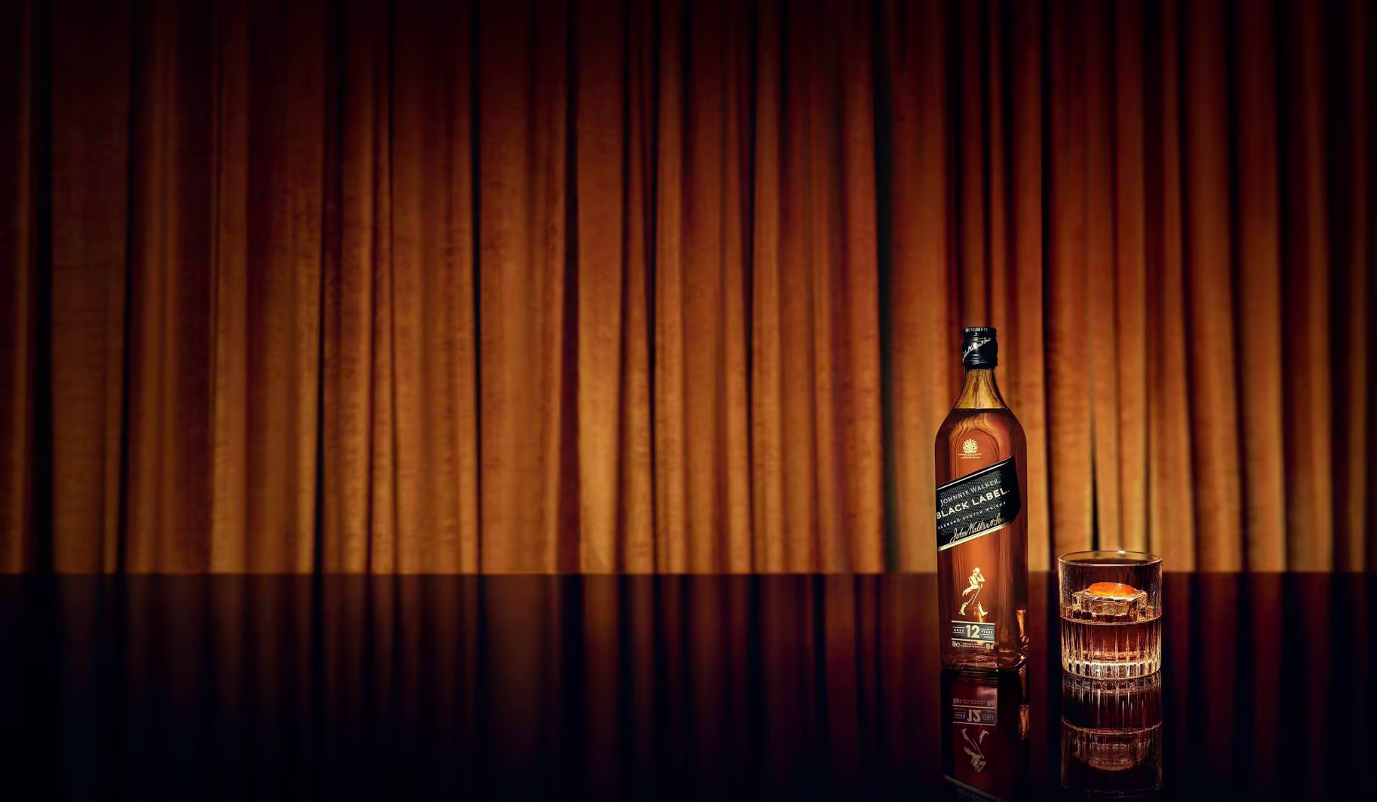 A bottle of Johnnie Walker Black Label and a tumbler of whisky on a reflective table in front of an amber curtain.