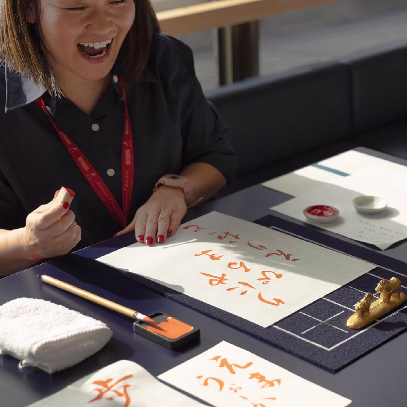 A woman smiles while practicing calligraphy with orange ink at a table set with writing tools and paper.