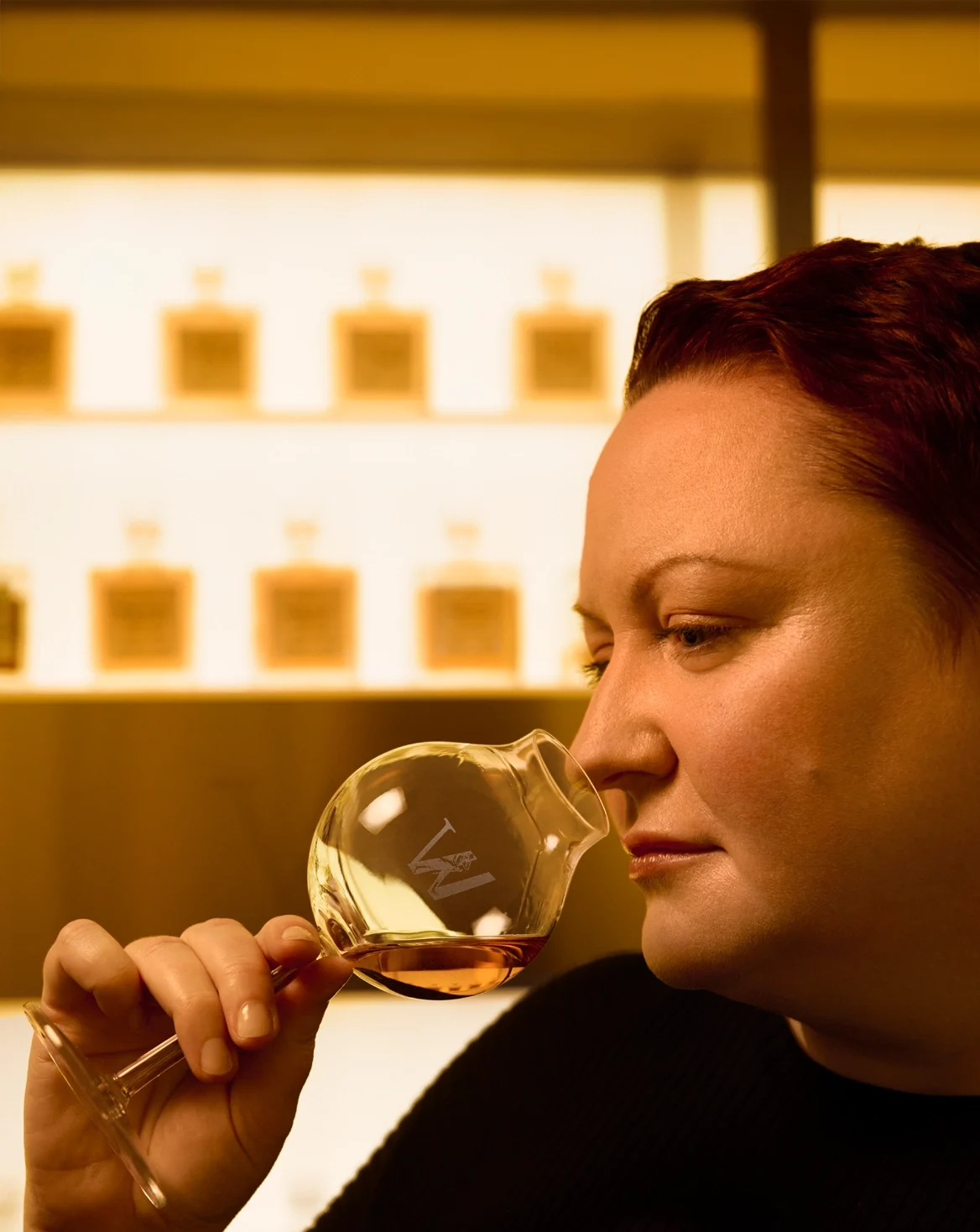 A person with red hair is holding and sniffing a glass of whiskey in a dimly lit room. Shelves with bottles are blurred in the background, creating a warm ambiance. The person appears thoughtful and concentrated on the aroma of the drink.