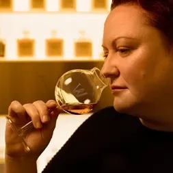 A person with red hair is holding and sniffing a glass of whiskey in a dimly lit room. Shelves with bottles are blurred in the background, creating a warm ambiance. The person appears thoughtful and concentrated on the aroma of the drink.