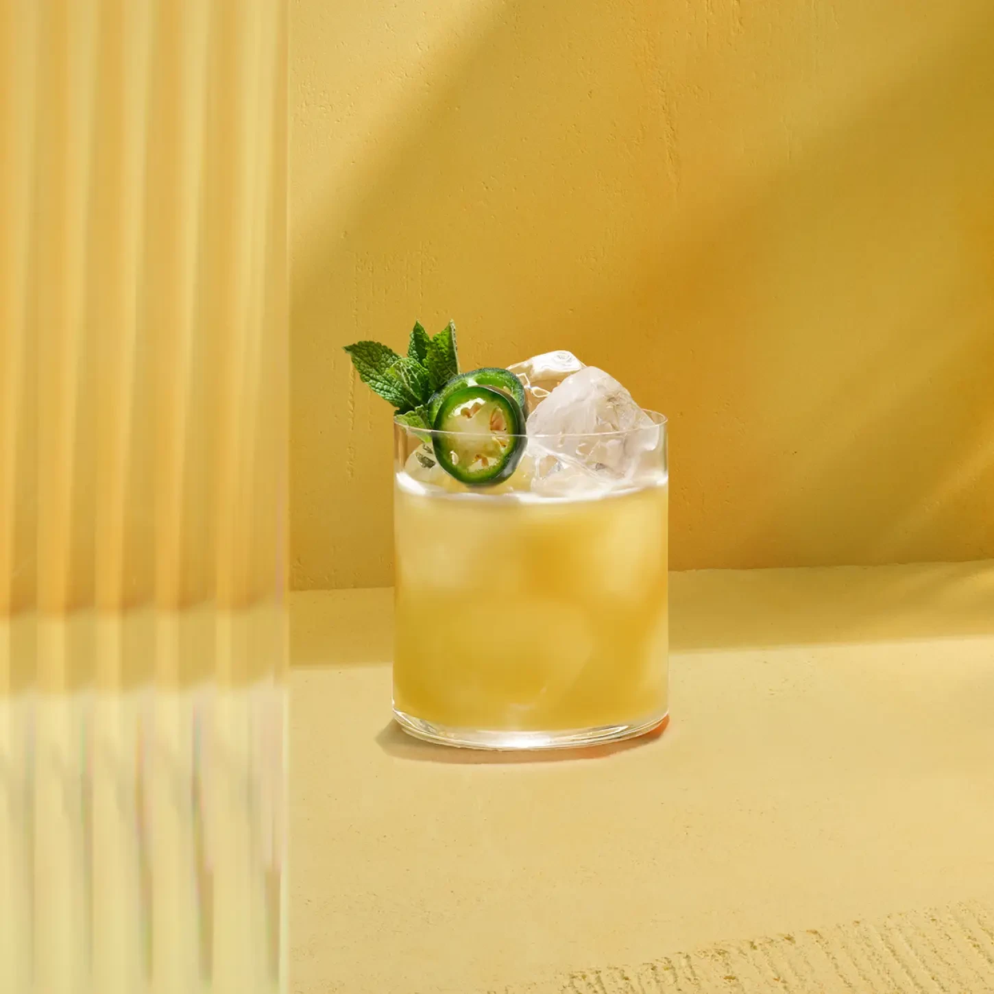 The Spicy Citrus Southside. A glass filled with a yellow cocktail on a sunny yellow table. The drink is garnished with ice cubes, a jalapeño slice, and a sprig of mint. Bright lighting casts reflections on the table.
