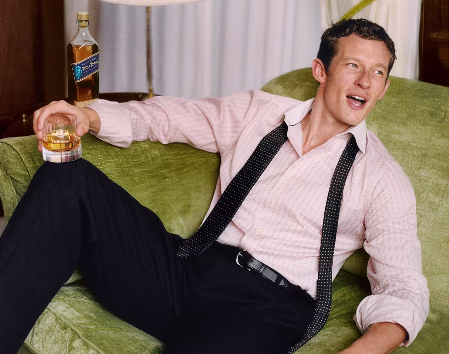 Callum Turner relaxing on a green couch, holding a glass of whiskey, bottle beside him, black tie undone.