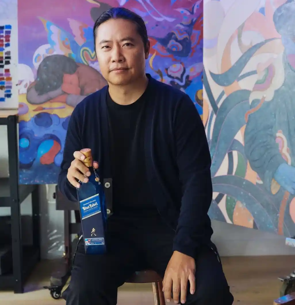 身穿深色服裝的 James Jean 手持一瓶 Johnnie Walker Blue Label 威士卡坐著。背景是一幅色彩繽紛的抽象畫，桌上放著藝術家的調色板。