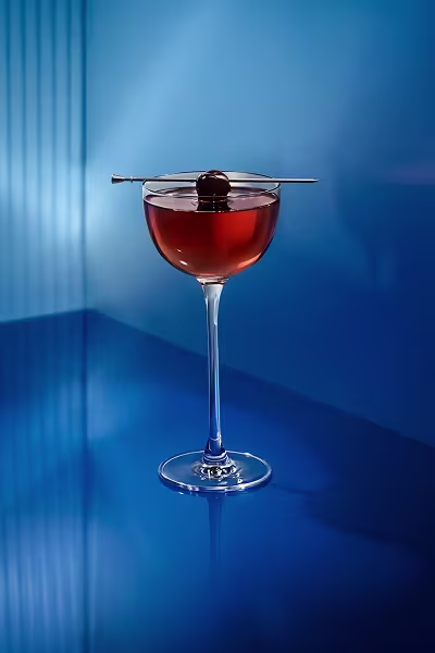 A cocktail in a coupe glass with a cherry garnish on a pick, set against a blue background.