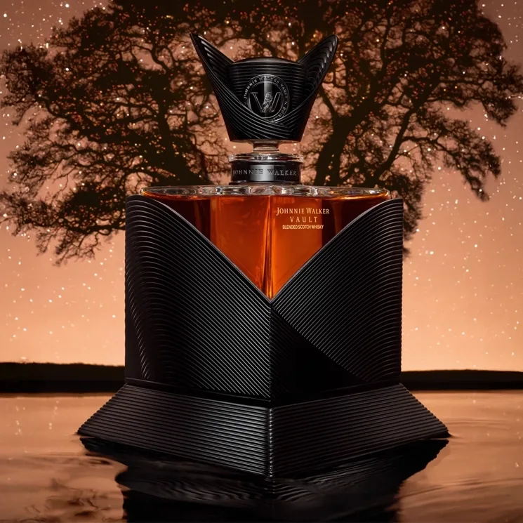 A luxurious bottle of Johnnie Walker whisky with an intricate black and gold geometric design is displayed in front of a large silhouetted tree and a softly glowing starry sky background.
