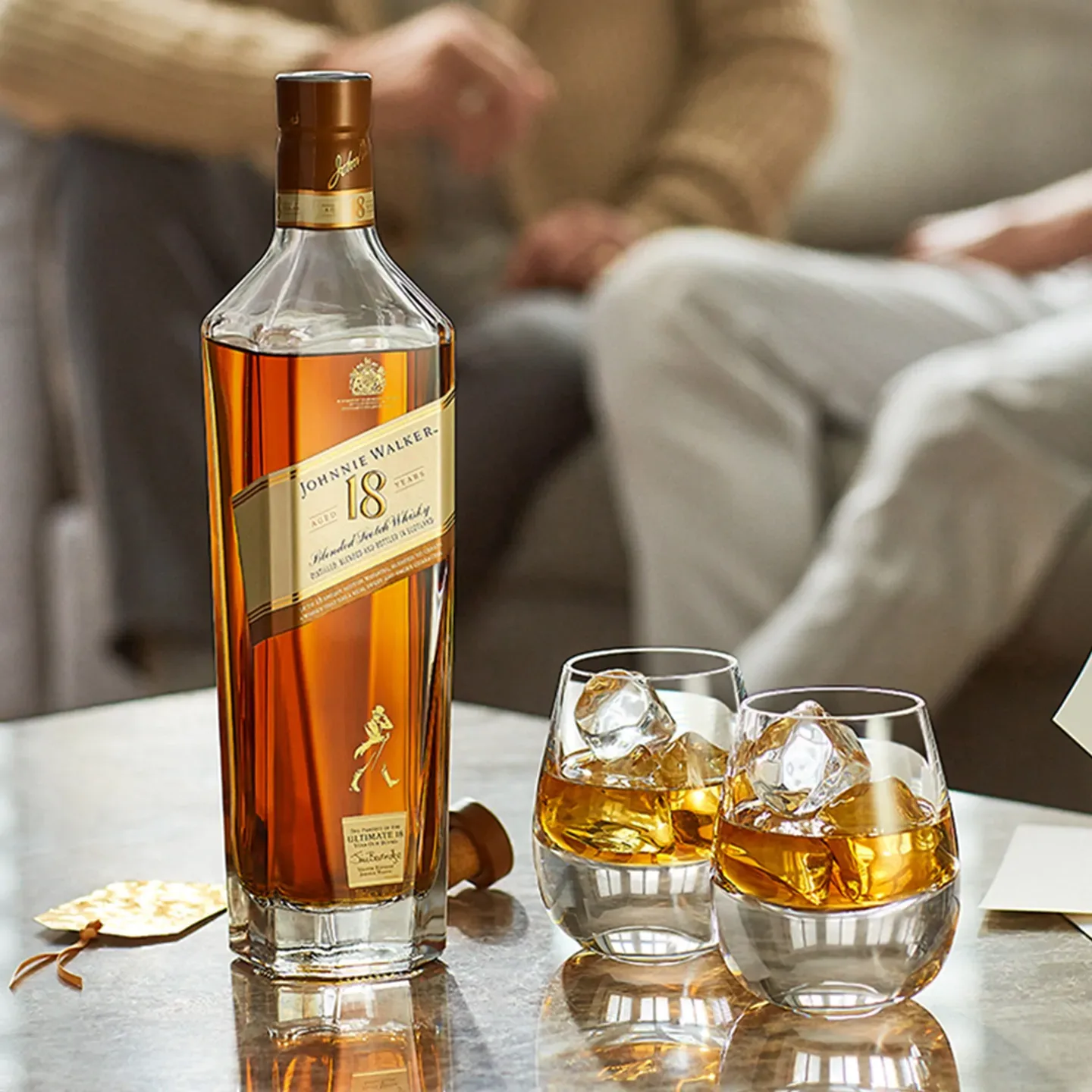 A bottle of Johnnie Walker 18-Year-Old Scotch sits on a table alongside two glasses filled with whisky and ice cubes. In the background, two people are partially visible, sitting on a couch.