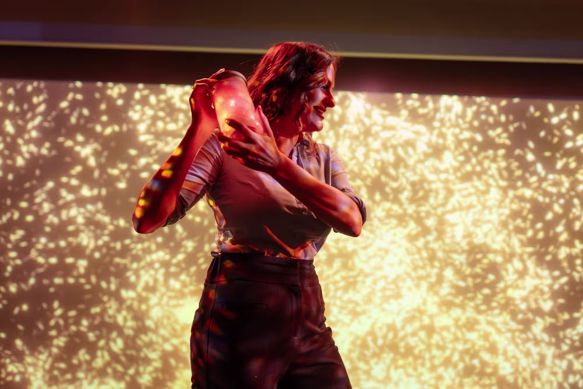 A woman smiles while shaking a cocktail shaker, standing in front of a bright, sparkling golden background that looks like fireworks or shimmering lights.