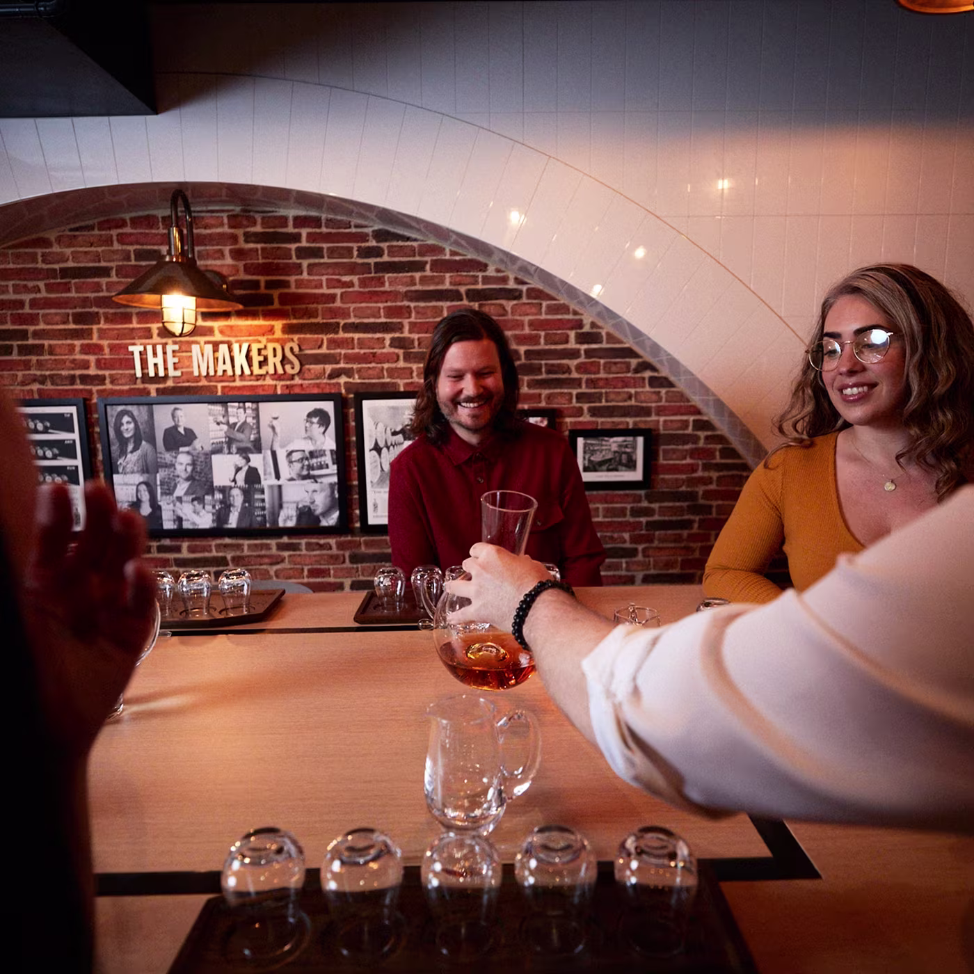 Three people gather at a bar with glasses; one pours a drink as others smile near a The Makers sign.