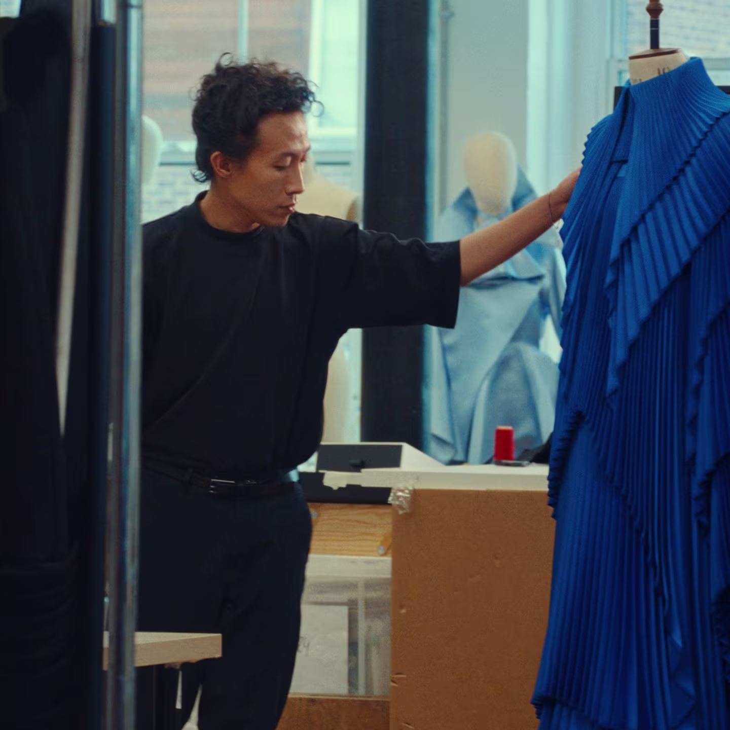 A person examines a blue pleated garment on a dress form in a fashion studio.