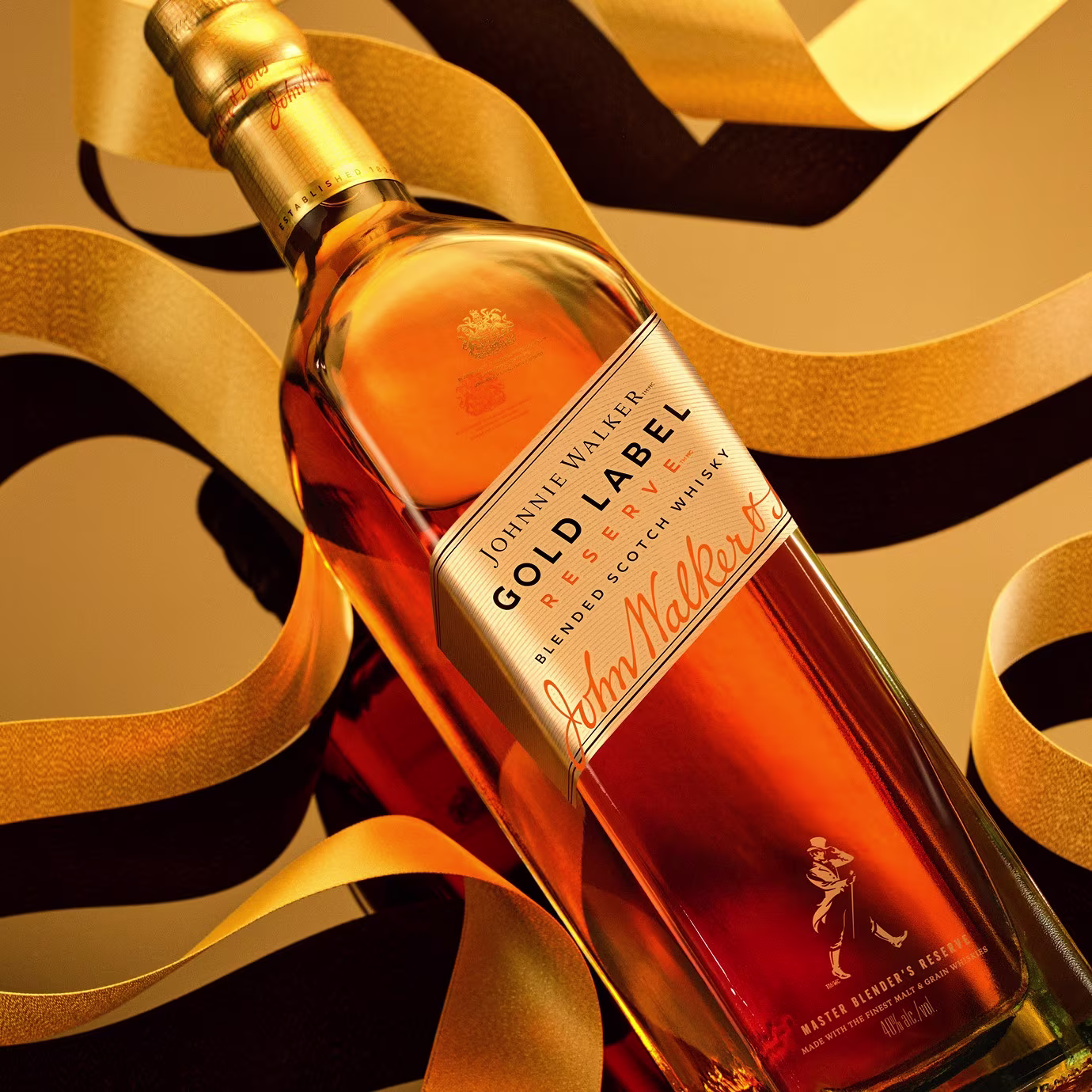 A bottle of Johnnie Walker Gold Label Reserve whisky lies on its side, surrounded by curling gold ribbons, against a warm golden background.