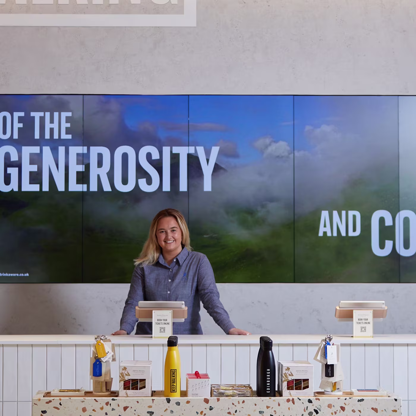 A woman stands smiling behind a counter with water bottles in front of a large screen displaying text.