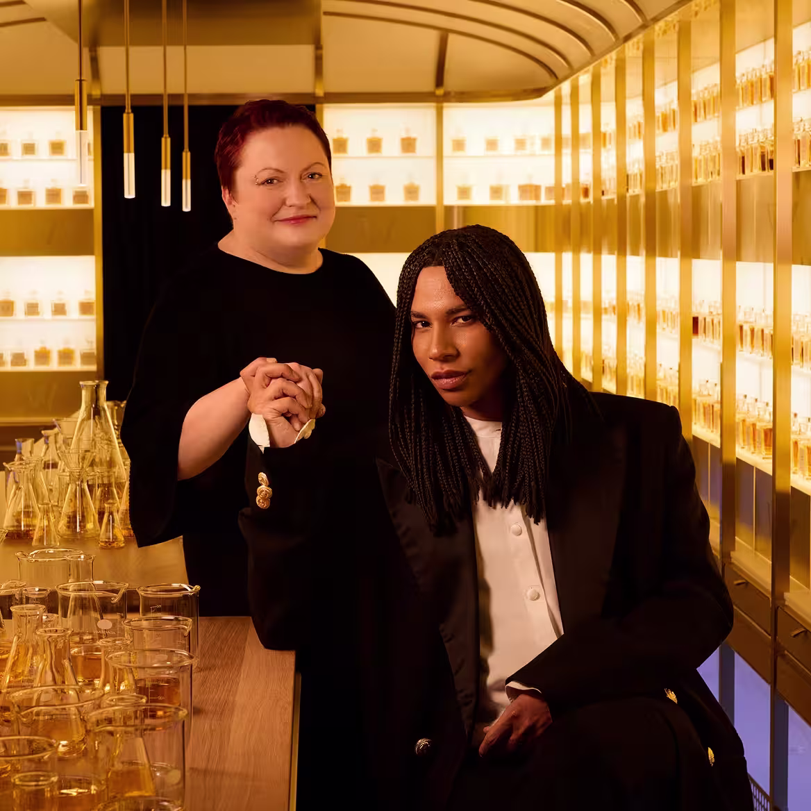Two people pose in a warmly lit room lined with perfume bottles. One stands smiling behind the other, who sits holding their hand, wearing a black jacket and white shirt. Glass vials and flasks with amber liquids are on the table.