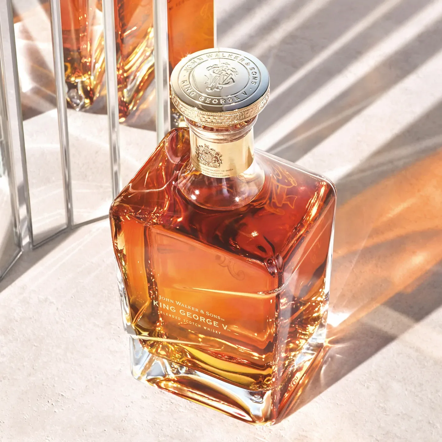 A square glass bottle of Johnnie Walker King George V whisky is filled with amber liquid and topped with a gold and cream cap. Sunlight casts shadows across the surface, highlighting the bottles elegant design.