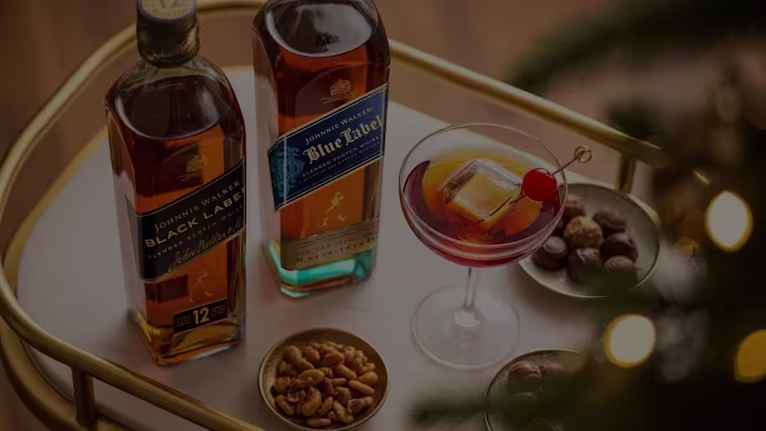 Twee flessen Johnnie Walker whisky, een glas cocktail met kersengarnituur en kleine schaaltjes met gemengde noten staan ​​op een ronde goud-witte serveerwagen met feestelijke verlichting op de achtergrond.