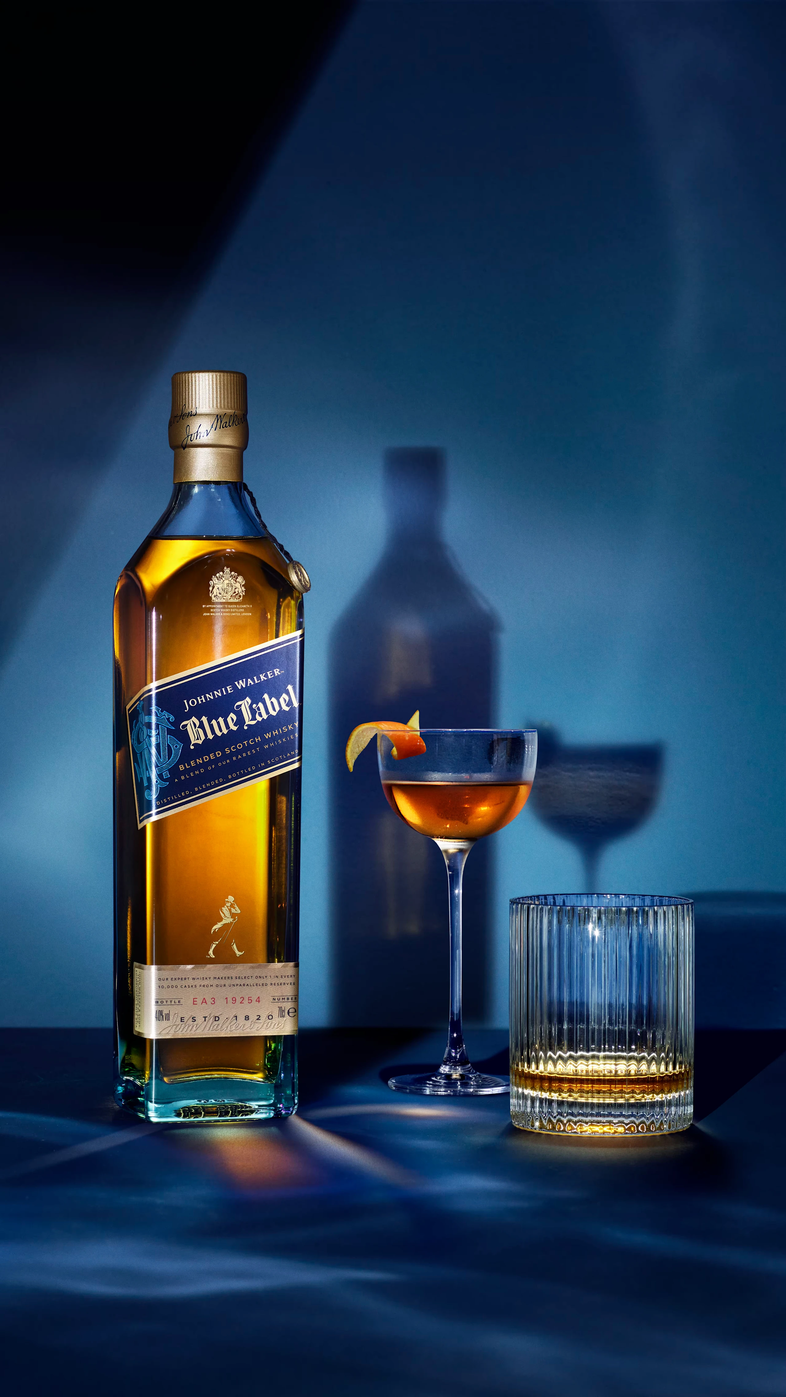 A bottle of Blue Label whiskey is displayed next to a cocktail glass with an orange peel garnish and a tumbler. The scene is set against a dark blue background with shadows reflecting the bottles silhouette.