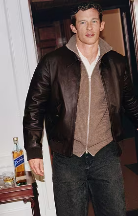 Cullum Turner in a brown leather jacket stands indoors by a table with whiskey, glasses, and a white wall behind him.