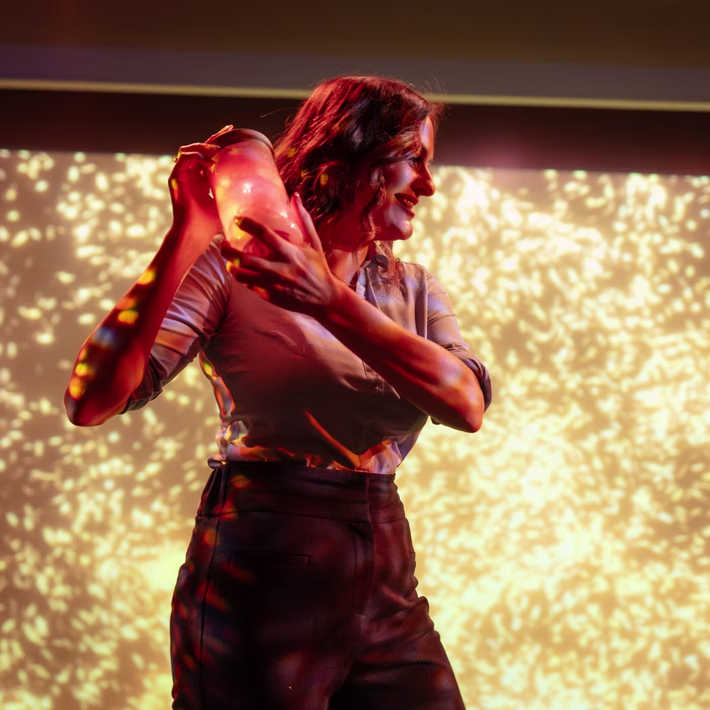 A woman smiles while shaking a cocktail shaker, illuminated by warm, dappled light patterns.