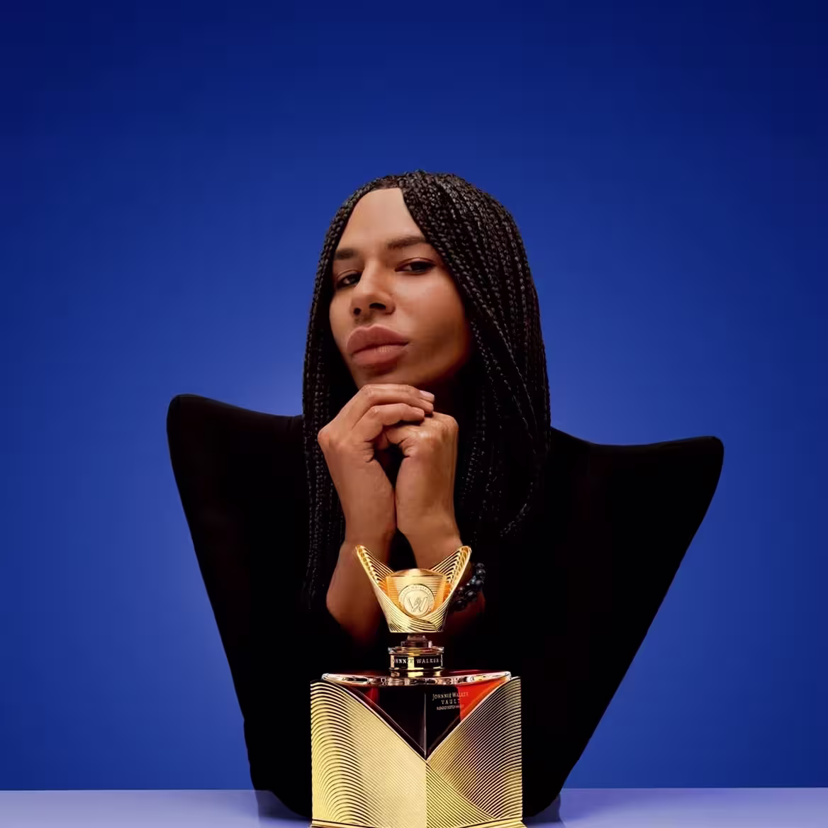 A person with braided hair and a serious expression poses with hands clasped under their chin, wearing a black outfit with angular shoulders. In front of them is an ornate, gold, geometric perfume bottle.