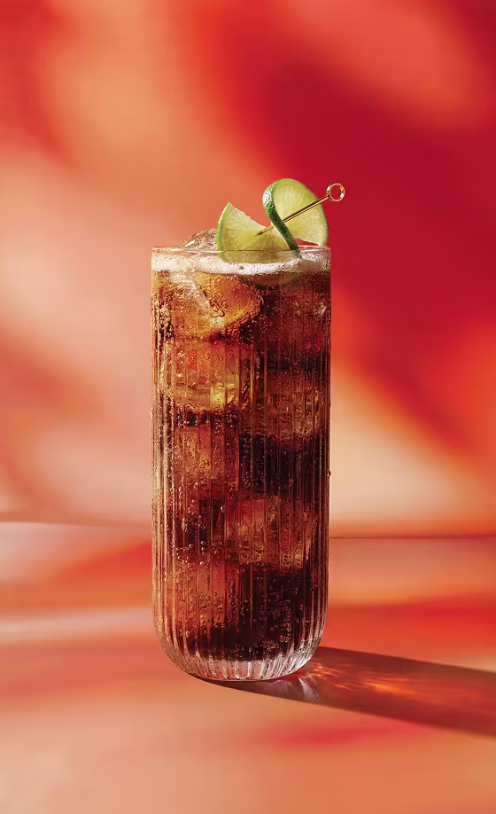 Ein hohes Glas Cola mit Eis und Limettenstücken vor einem warmen, orange-roten, verschwommenen Hintergrund.