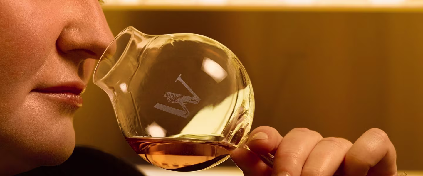 A person holding a round glass of amber liquid close to their nose, appearing to smell it. The glass has a W etched on it, with a blurred golden background.