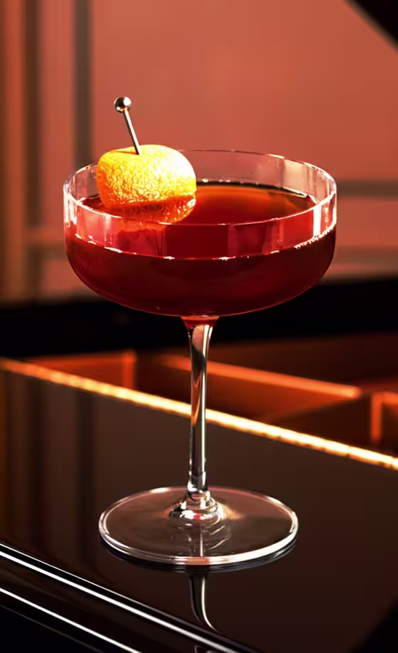 A deep red cocktail garnished with an orange peel sits in a coupe glass on a glossy black surface, reflecting warm, ambient lighting.