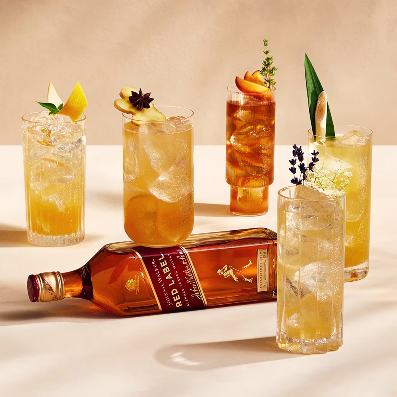 A bottle of Johnnie Walker Red Label and five garnished cocktails on a cream-colored surface.