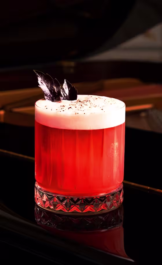 A vibrant red cocktail with a frothy top, garnished with dark leaves, sits in a textured glass on a glossy black piano. Soft light highlights the drink’s rich color and elegant presentation.