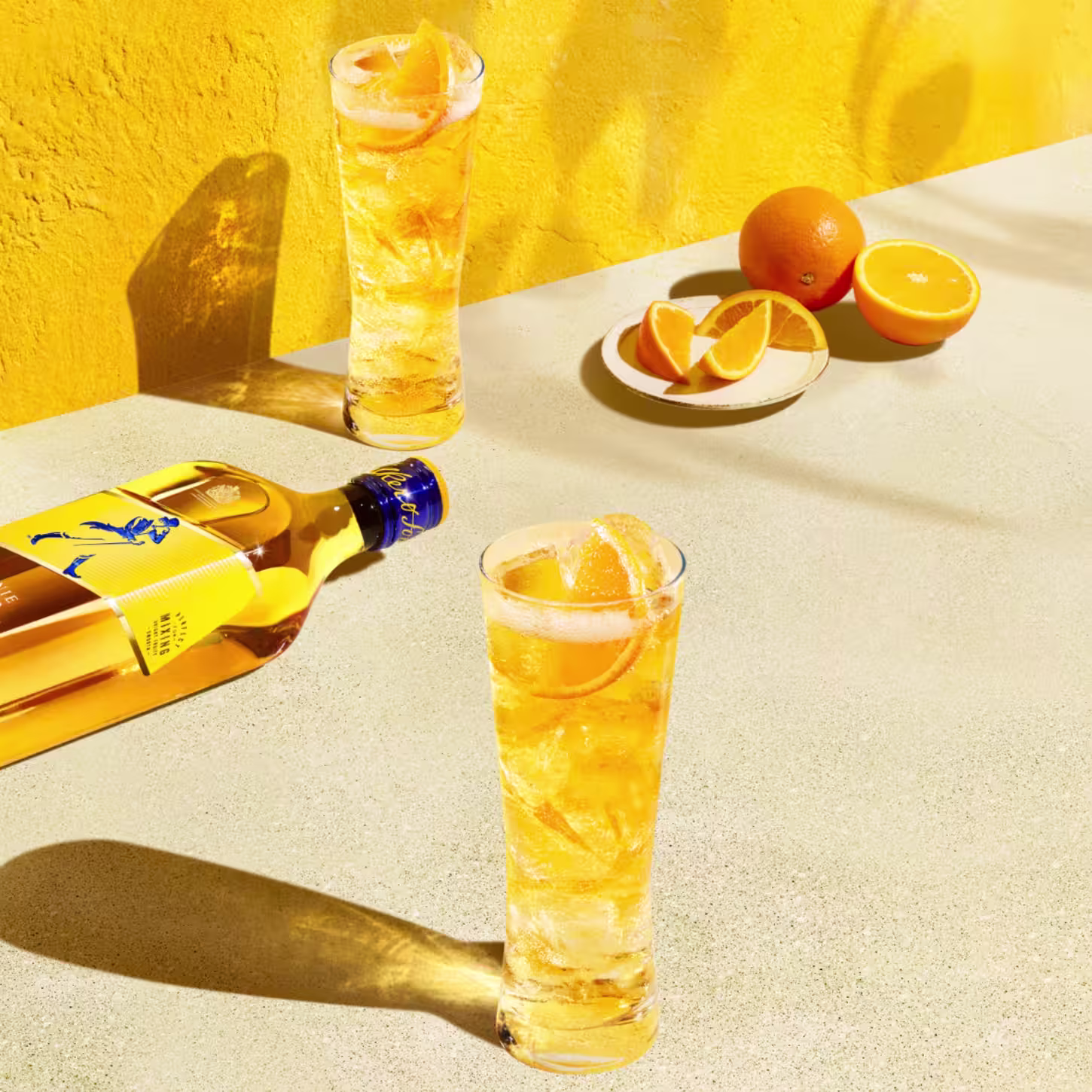 兩個高腳玻璃杯,裝滿金色飲料,飾有橙片,放在陽光照射的表面上。附近有一個威士忌酒瓶,背景是一堵充滿活力的黃色牆壁,上面放著一個裝有橙片的盤子。