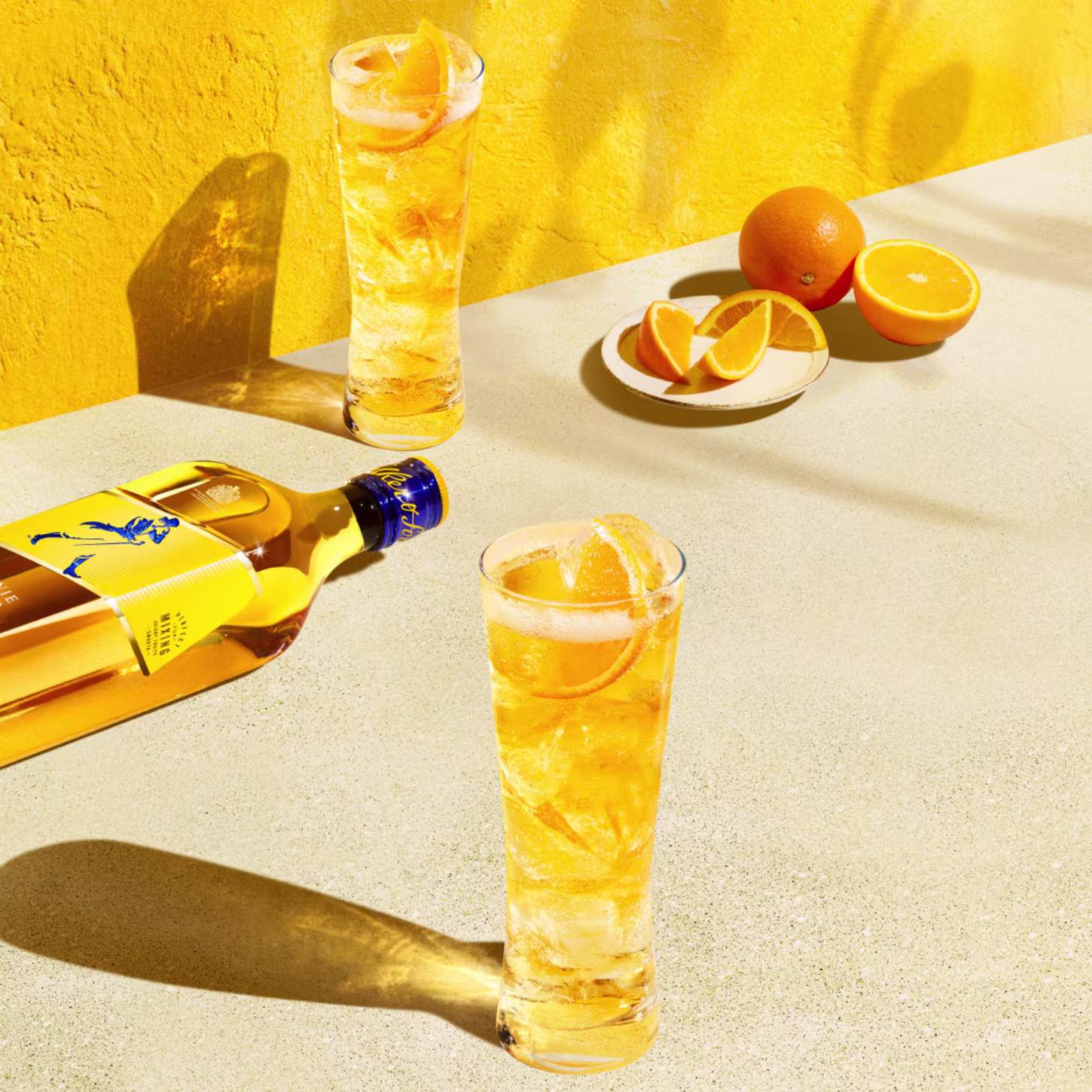 Two tall glasses filled with a golden drink garnished with orange slices, placed on a sunlit surface. A whiskey bottle lies nearby, and a plate with orange slices is in the background against a vibrant yellow wall.