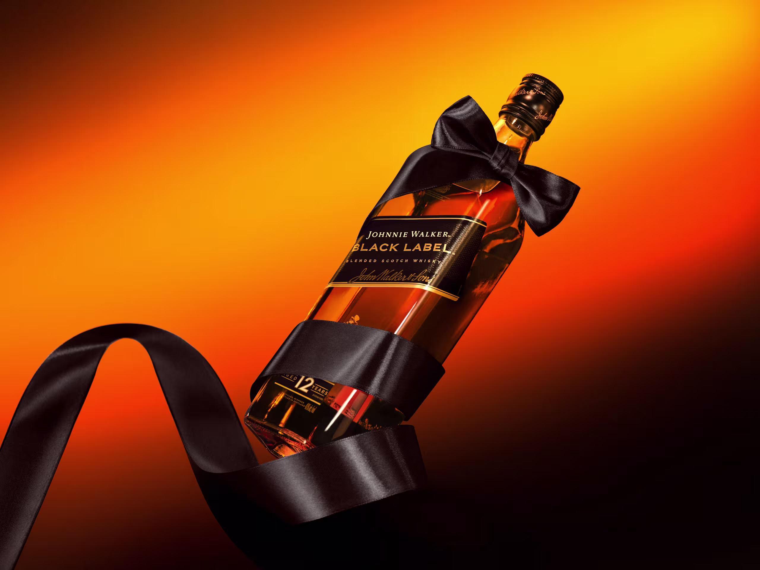 Een fles Johnnie Walker Black Label whisky is verpakt met een glanzend zwart lint en strik, tegen een opvallende oranje en gele achtergrond.