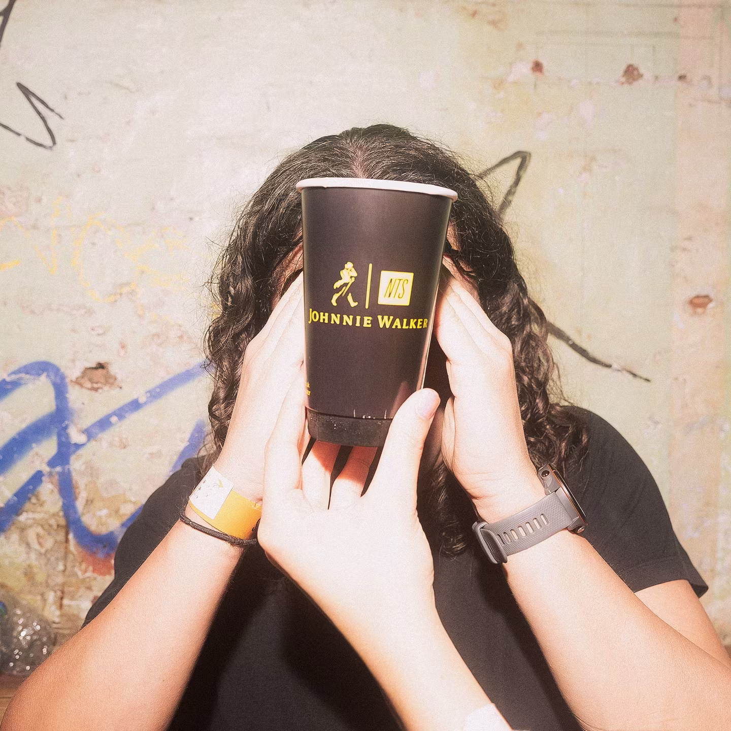 Person covering their face with a Johnnie Walker branded cup, wearing a wristband and watch, against a graffiti wall.