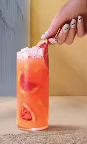 A hand with painted nails garnishes a tall glass of orange-pink drink with sliced citrus and crushed ice, set against a modern, colorful background.