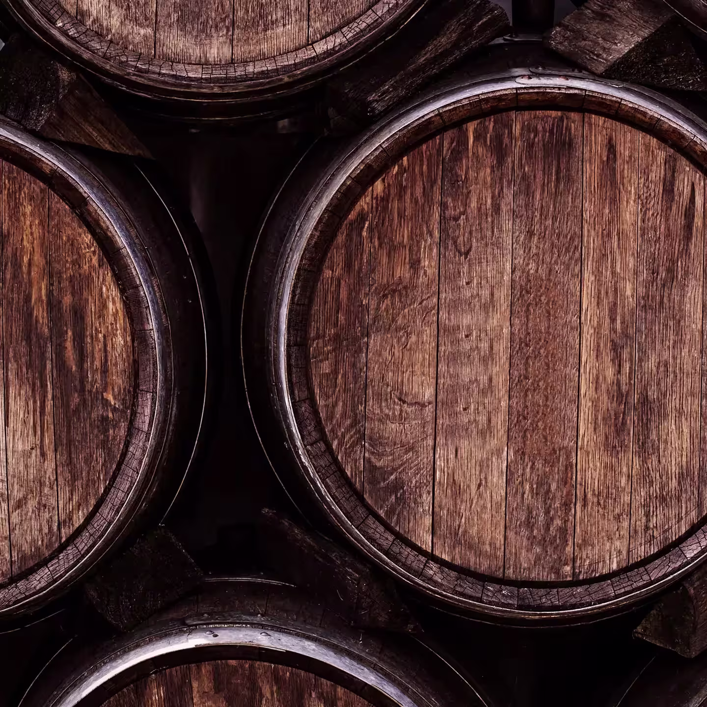 Close-up of wooden barrels arranged horizontally, showcasing the weathered texture and grain of the wood. The barrels, likely used for aging beverages like whiskey or wine, are stacked in a dimly lit setting.