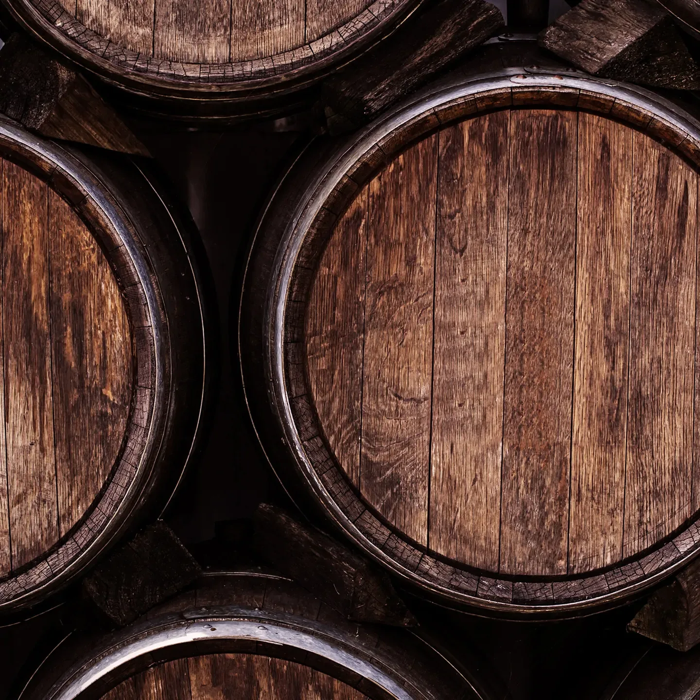 Close-up of wooden barrels arranged horizontally, showcasing the weathered texture and grain of the wood. The barrels, likely used for aging beverages like whiskey or wine, are stacked in a dimly lit setting.