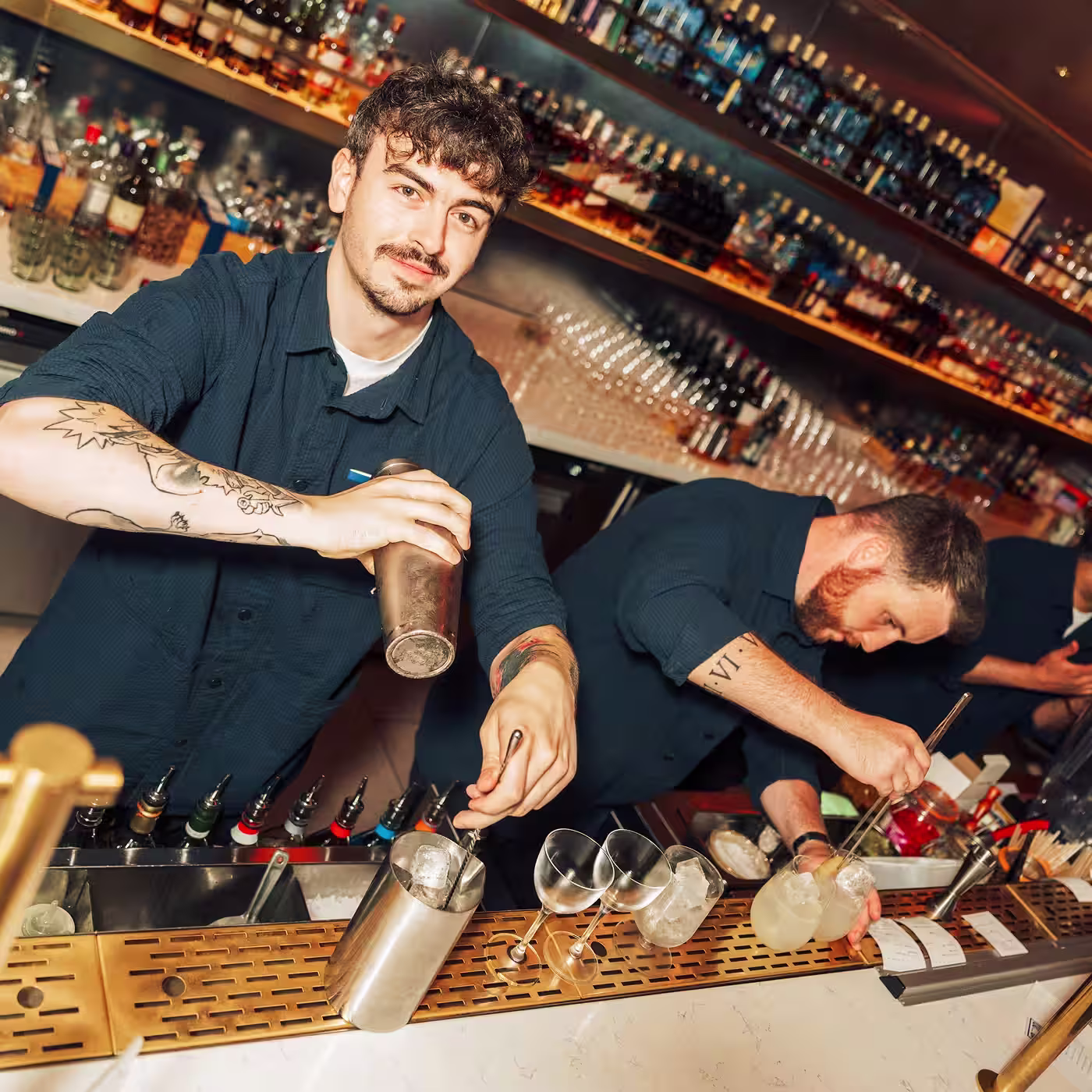 Two bartenders mixing drinks behind a bar, one smiling at the camera while shaking a cocktail.