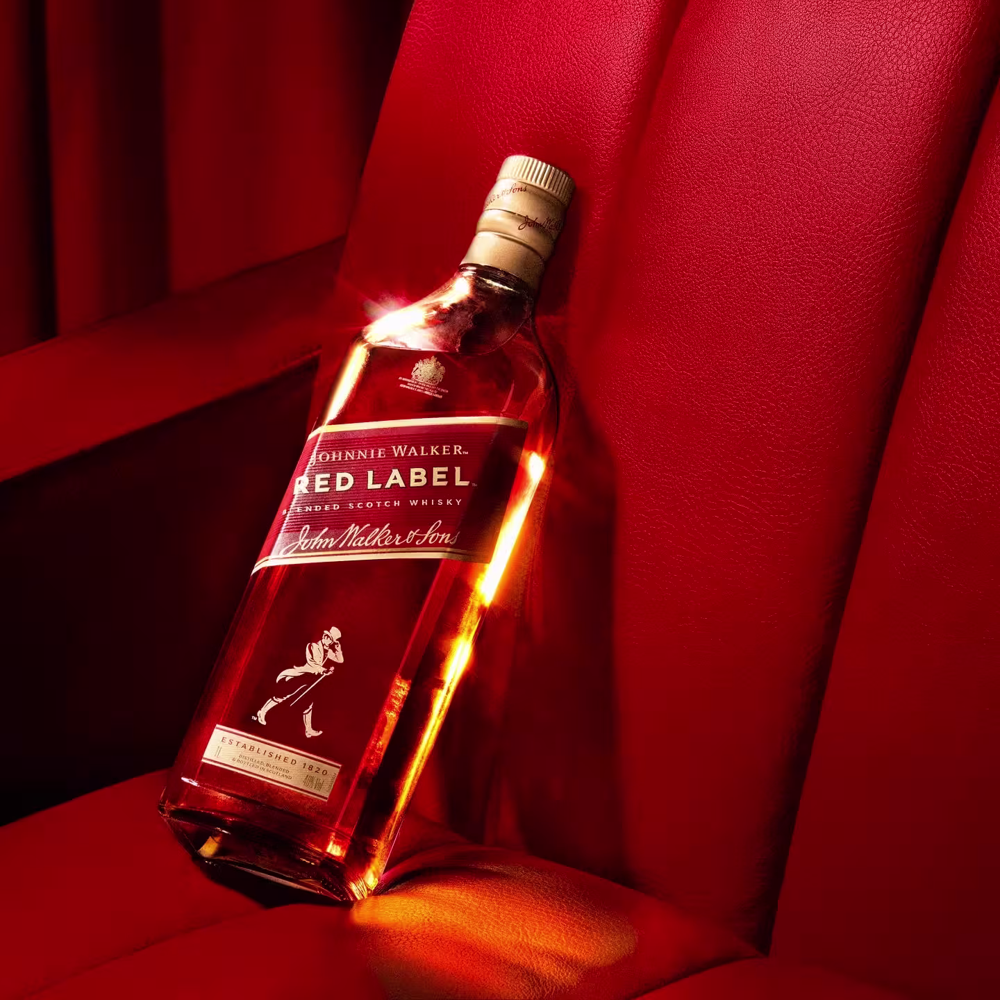 Una botella de whisky Johnnie Walker Red Label descansa sobre el respaldo de un asiento de cuero rojo, iluminado por una luz cálida.