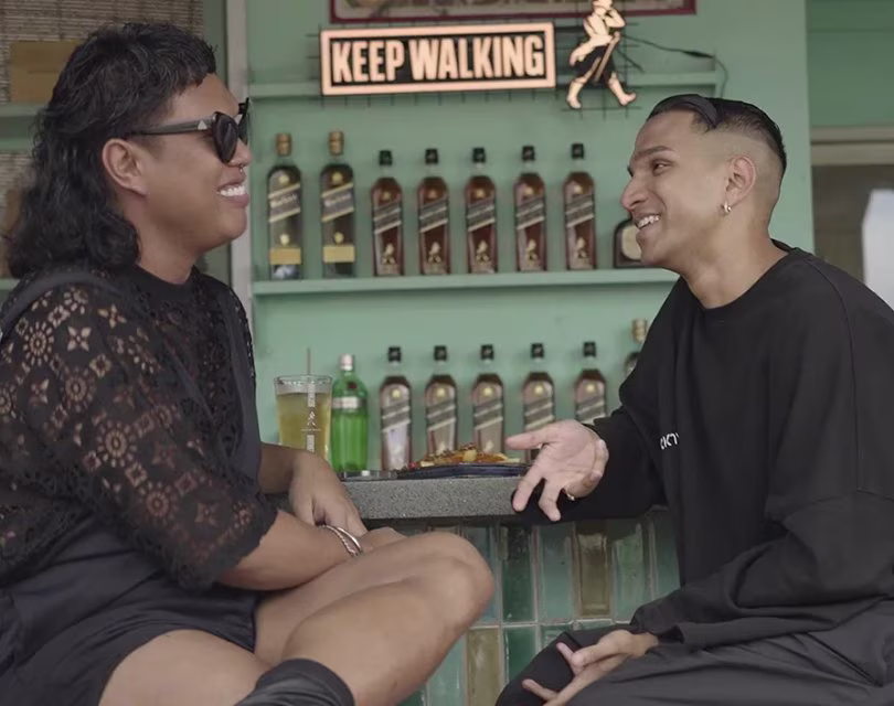 Two people sit and chat at a bar with bottles displayed behind them. They are smiling and appear to be enjoying their conversation. A sign above them reads KEEP WALKING.