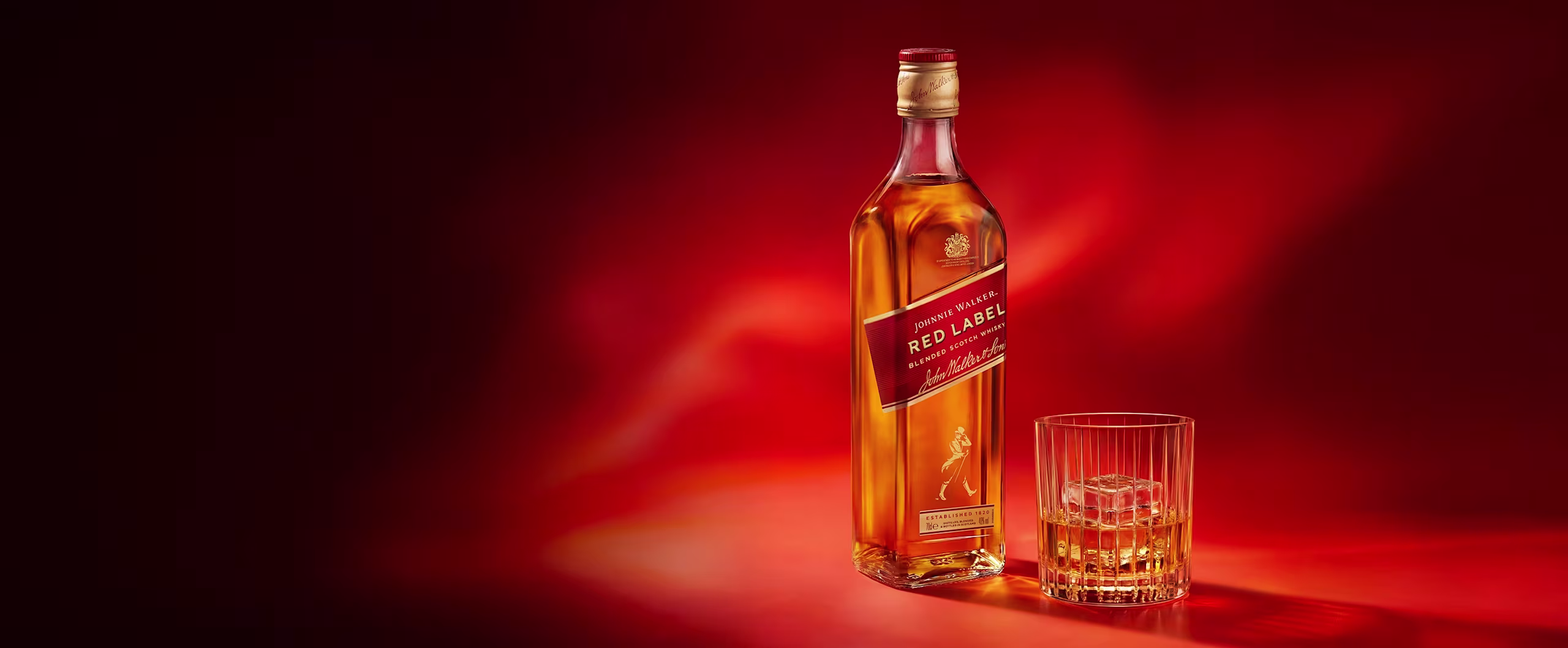 Una botella de whisky Johnnie Walker Red Label junto a un vaso con whisky y hielo sobre un fondo rojo.