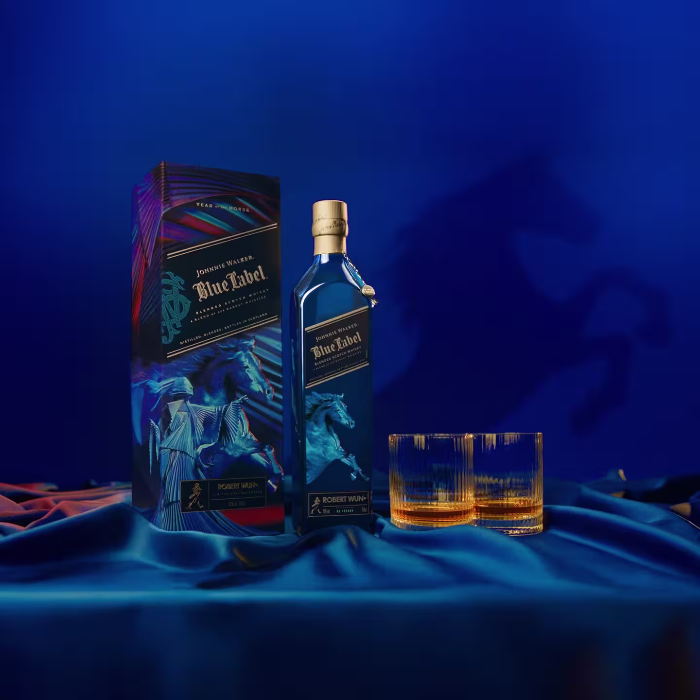 A Johnnie Walker Blue Label whiskey bottle and decorative box sit on a blue and orange fabric with two empty glasses nearby; a blue horse silhouette is projected in the background.